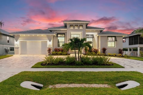 A contemporary single-story home with tropical landscaping and a two-car garage, captured during a stunning sunset with pink and purple hues filling the evening sky.