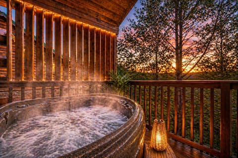 Private hot tub with mountain views at sunset, featuring warm string lights, glowing lantern, and relaxing steam. A cozy, romantic cabin deck perfect for couples looking to unwind in a peaceful forest setting.