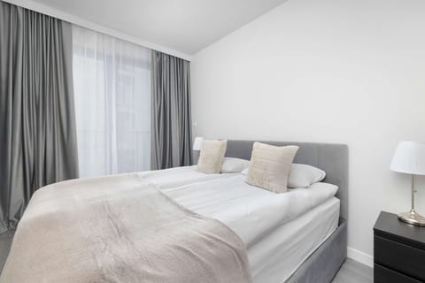 A bright bedroom with a large double bed, white bedding, and soft grey curtains framing the window. A small bedside table with a lamp completes the simple, elegant arrangement.