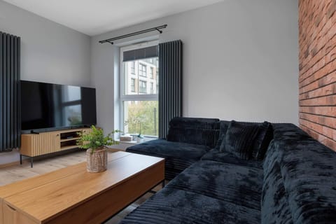 A bright living room features a dark sectional sofa paired with a light wood coffee table. An exposed red brick accent wall contrasts nicely with the modern TV stand and grey walls. Large windows let in plenty of natural daylight.
