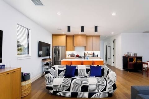 This modern open-concept space seamlessly blends living room and kitchen areas. Features include a gray sectional sofa, wall-mounted TV, unique zigzag bookshelf, and honey-toned wooden cabinetry with white countertops and stainless appliances.