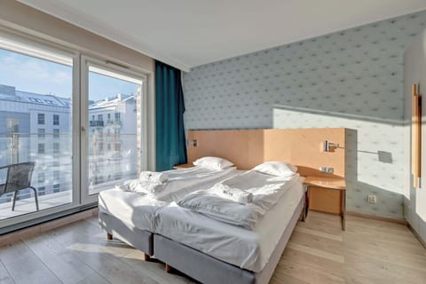 A bright bedroom with a double bed covered in white linens. A large window provides natural light and a view, while the modern decor includes light wood and soft colors.
