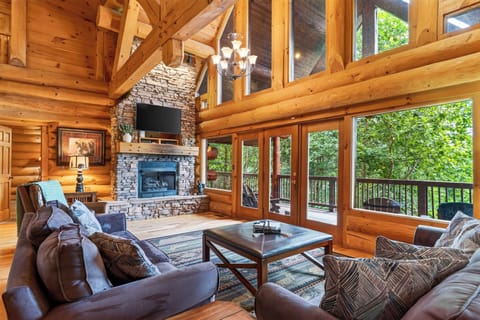 Soaring windows and a stone fireplace anchor this warm log-cabin living room, with direct deck access and leafy forest views just beyond.