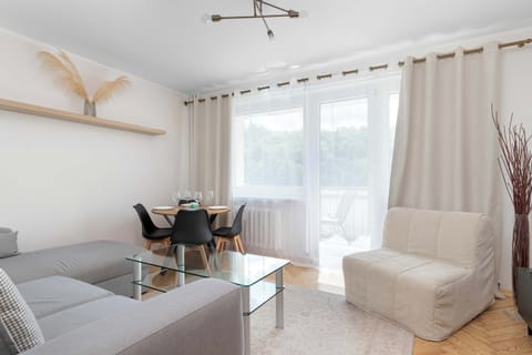 A bright room features a comfortable grey sofa and a glass coffee table. In the background, a dining table sits near a large window draped with curtains.