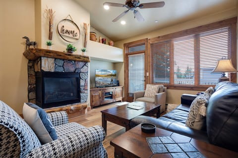 Settle into your cozy living room featuring a rustic stone fireplace, plush seating, and a ceiling fan. Sunlight filters through wood blinds, inviting you to unwind.
