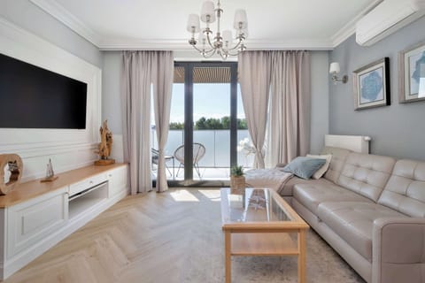 A bright living room with a sofa, coffee table, TV, and large windows leading to a balcony. The interior is decorated in a modern, neutral style.