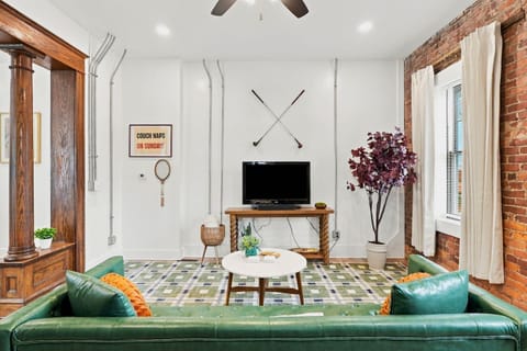 Stylish living room with plush green sofa, television, and historic exposed brick. Minutes from Promenade Park, Science Central, and local dining.