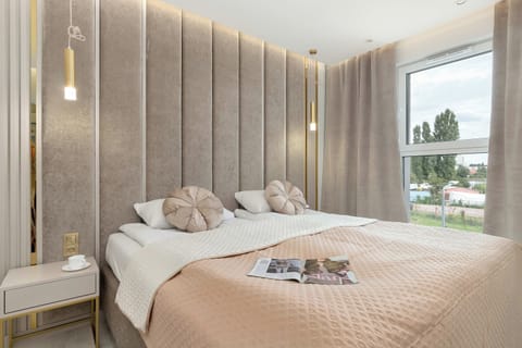 A cozy bedroom with a large upholstered bed, soft neutral bedding, and a textured wood-paneled accent wall. Floor-to-ceiling windows overlook the surrounding area.