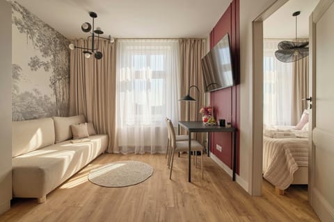 Stylowy i komfortowy salon z wygodną sofą, dużymi oknami i elegancką strefą wypoczynku. Ciepła drewniana podłoga, dekoracyjna tapeta, burgundowe akcenty i nastrojowe oświetlenie nadają wnętrzu charakter.