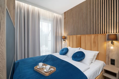 A stylish bedroom area with a large bed dressed in white and blue bedding, complemented by a warm wood slat accent wall. Floor-to-ceiling windows bring in plenty of natural light.