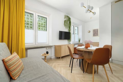A bright living area features a grey sofa bed and a dining table with chairs. Sunlight streams through a window accented with yellow curtains.