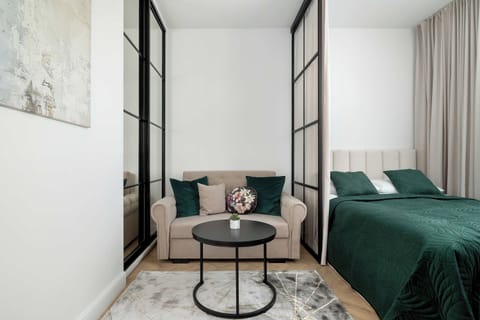 A cozy bedroom with a green bed, armchair, round coffee table, and large windows.