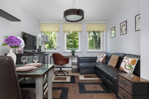 A bright living room with a dark sofa, armchairs, a dining table, and colorful accents, illuminated by large windows.