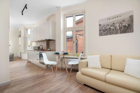 An open-concept living space features light wood-look flooring and an elongated leather sofa positioned against the wall. Large vertical windows let in natural light, leading the eye toward the dining table and kitchenette in the background.