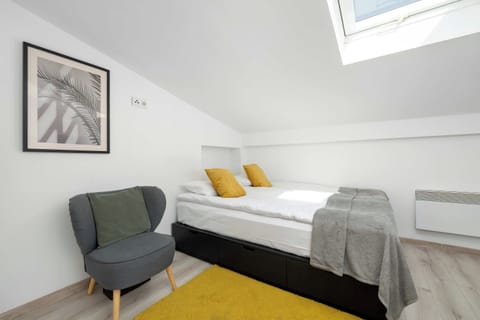 A bright bedroom featuring a double bed with a gray runner and two yellow pillows. A comfortable gray armchair sits next to a yellow rug under a framed picture on the wall. A skylight lets natural light into the sloped-ceiling room.