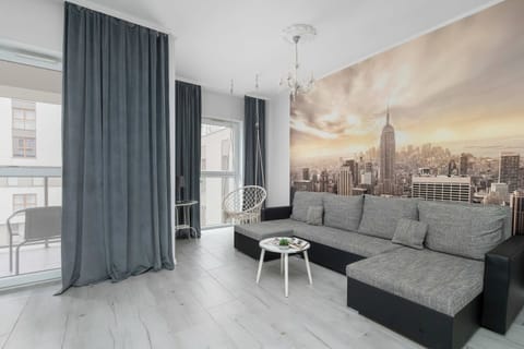 A modern living room with a large gray sectional sofa, a small round coffee table, and a wall-mounted TV. Floor-to-ceiling curtains and large windows provide plenty of natural light.