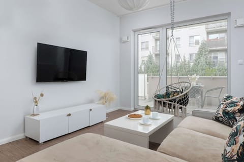 A bright living room featuring a comfortable beige sofa and a flat-screen TV on a white stand. A dining area is visible in the background by the window.