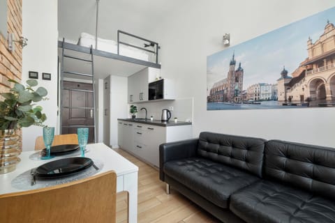 A bright studio with a black leather sofa and a compact dining area. The space features a well-equipped kitchenette, wooden flooring, and decorative wall art.