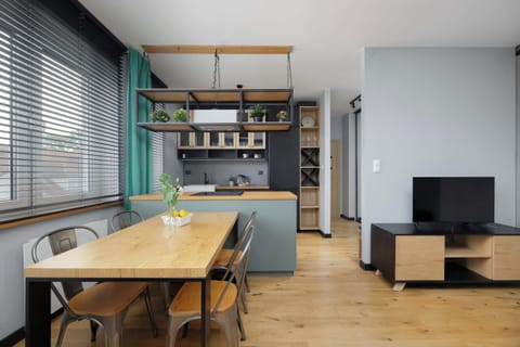 A spacious living area featuring a light wooden dining table and matching chairs. A modern black television rests on a low console against a smooth gray accent wall.