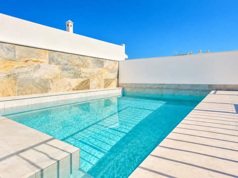 Private turquoise swimming pool at a modern beach villa, featuring elegant stone accents, clean white walls, and a sun-drenched deck under a clear blue sky.