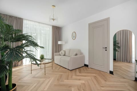 A wide shot of an apartment interior featuring a living area with a light sofa, wooden floors, and a view into a hallway. Large windows let in plenty of daylight to brighten the space.