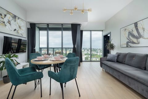 Open-plan living space featuring a grey sofa and a dining area. Dark green chairs surround a wooden table near large bright windows.