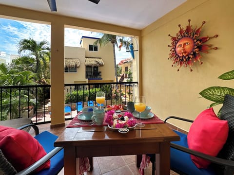 A2-10-2026 Private Balcony - Private balcony at Abbey del Sol A2 Unit 10, a Puerto Morelos vacation rental, featuring a table with colorful place settings, red cushions, tropical plants, and a view over the pool and courtyard.