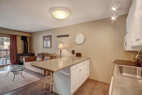 A cozy living room and kitchen area features a small sofa, wall clock, wood paneling, and modern lighting fixtures. A glass coffee table and a wood-burning stove add to the room's comfort and functionality.