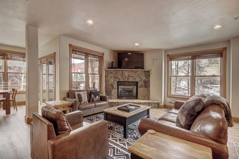 A cozy living room with leather sofas, a stone fireplace, a mounted TV above it, and large windows letting in natural light. The space is decorated in a rustic style with wooden furniture and accents.