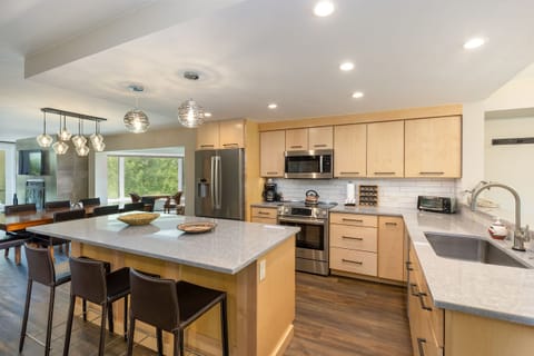 A modern kitchen with light wood cabinets, stainless steel appliances, a large island with seating for three, and a view into a dining area with a table and chairs next to large windows.