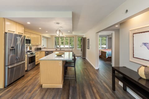 A modern kitchen with wooden cabinets, stainless steel appliances, an island with bar stools, and adjacent dining and living areas with hardwood floors and large windows.
