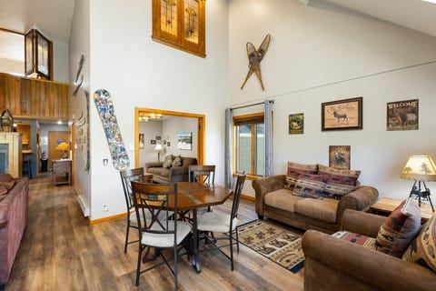A cozy living and dining area featuring a round table with four chairs, two brown sofas, a wall-mounted snowboard, and various rustic decor and signs. A staircase leads to an upper loft.