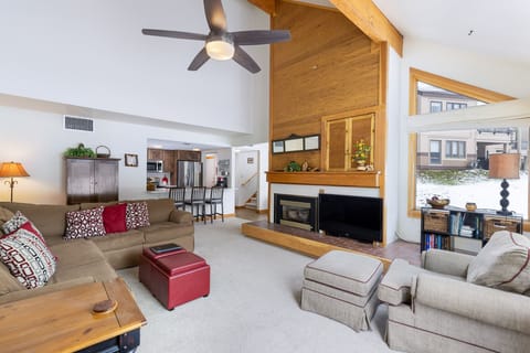 A spacious living room features a large ceiling fan, a sectional sofa, a fireplace, and a flat-screen TV. The room includes wooden accents, high ceilings, and windows with a snowy exterior view.