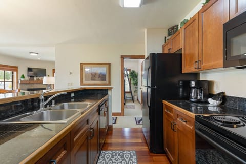 A modern kitchen with wood cabinets, black countertops, and appliances including a refrigerator and microwave. A double sink is present on the island. The room is well-lit with a ceiling light.