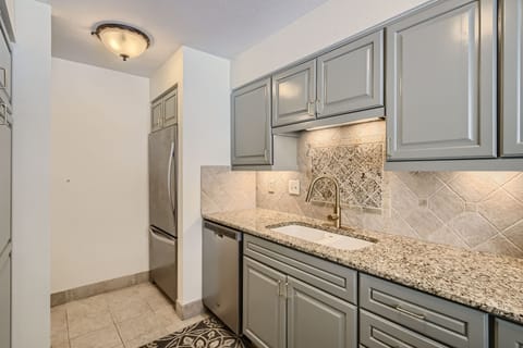 A modern kitchen with grey cabinets, granite countertops, stainless steel fridge, under-cabinet lighting, and a tiled backsplash above a white sink with a sleek brass faucet.