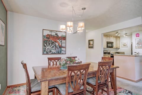 A dining room with a wooden table and six chairs, an overhead light fixture, wall art featuring a red tractor, and a view of a kitchen with light-colored cabinetry and appliances.