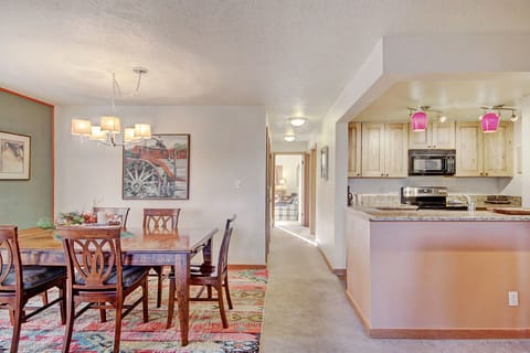 A dining area with a wooden table set for six next to a kitchen. The kitchen has light wood cabinets, granite countertops, and pink pendant lights. There's a hallway leading to other rooms.