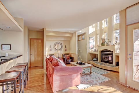 A cozy living room with a red couch, wooden floors, a fireplace, bar seating, and large windows letting in natural light.