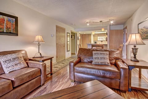 A cozy living room with brown leather sofas, wooden furniture, and a view into the kitchen with a bar counter and stools. Walls decorated with landscape art and warmly lit lamps.