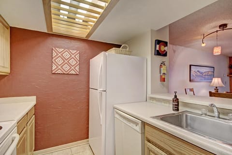A compact kitchen features a white refrigerator, dishwasher, and sink, with a red accent wall and decorative canvas. A basket sits on top of the fridge, and a fire extinguisher is mounted nearby.