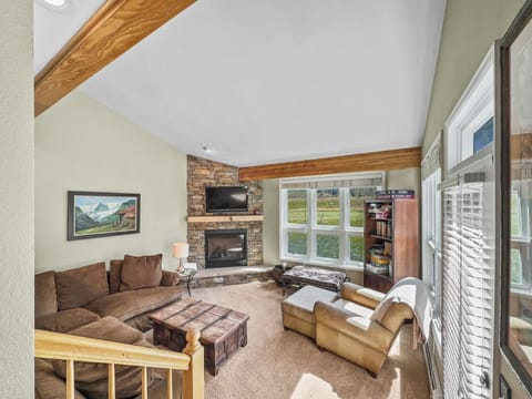 A living room with a brown sectional sofa, a stone fireplace, a wooden coffee table, and a bookshelf. The room features a large window and exposed wooden beams on a high ceiling.