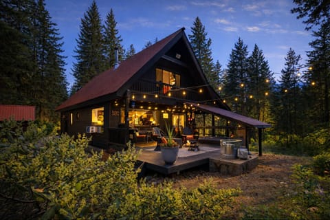 A stunning forest cabin with warm lighting and outdoor deck, nestled among towering evergreens at twilight.