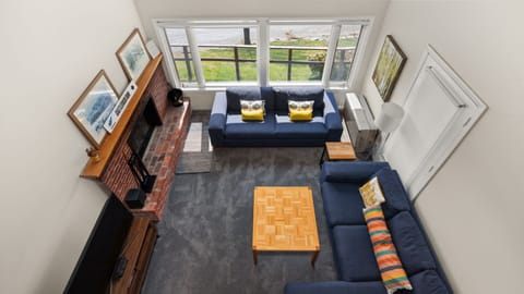 A bird's-eye view of a living room with two blue sofas, a fireplace, and a wooden coffee table. Large windows offer an outside view, and the room has framed artwork and a floor lamp.