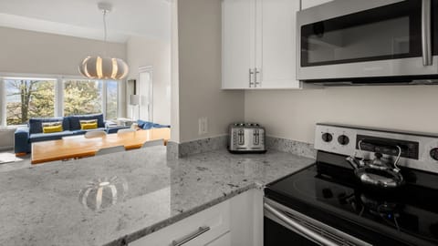A modern kitchen with gray countertops, a stainless steel stove, a microwave, a toaster, and a teapot. The background shows a dining area with a wooden table, chairs, and a large window.