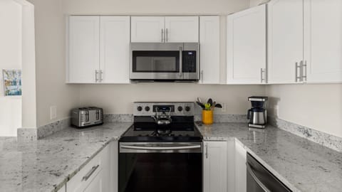 Modern kitchen with white cabinets, stainless steel appliances including a microwave, stove, toaster, and coffee maker. A yellow utensil holder and a kettle are on the counter.