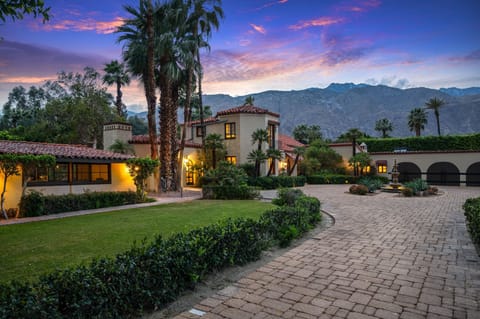 Spanish-style estate nestled against dramatic mountain backdrop, surrounded by lush palms and landscaped grounds, showcasing a welcoming entrance at twilight.