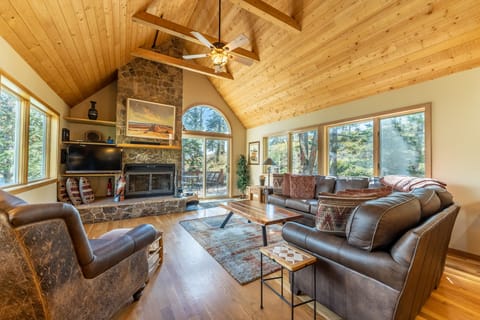 Unwind in your spacious living room featuring soaring wooden ceilings, cozy stone fireplace, and panoramic forest views through floor-to-ceiling windows.