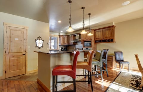 A modern kitchen with wooden cabinets and a central island with colorful bar stools. Three pendant lights hang above the island, and the room includes a microwave, stove, and wooden door.