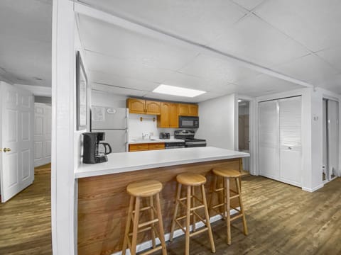 A small kitchen with wooden cabinets, countertop with three wooden stools, coffee maker, stove, microwave, and refrigerator. The floor is wooden, and there's a white door in the background.