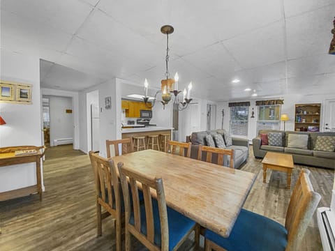 A modern, open-concept dining and living area featuring a wooden table with six chairs, a gray sofa, a wooden coffee table, and a kitchen with a breakfast bar. Chandelier lighting hangs above the table.